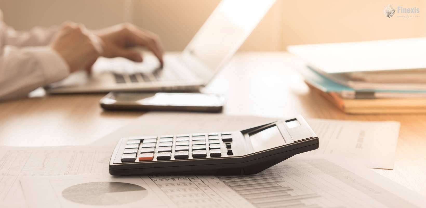 Zbliżenie na kalkulator leżący na dokumentach finansowych na biurku, w tle osoba pracująca na laptopie w jasnym biurze.jpg